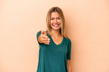 Young caucasian woman isolated on beige background stretching hand at camera in greeting gesture.