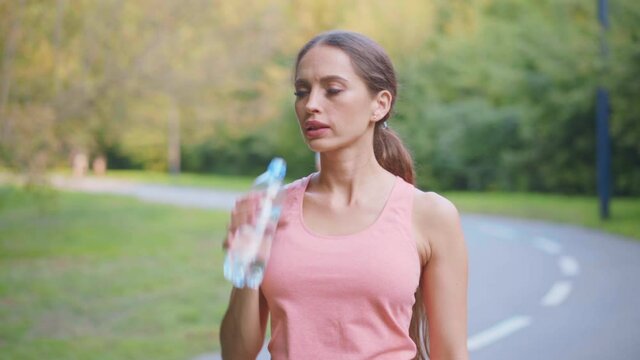 Female runner drink water transparent plastic bottle after morning jogging Caucasian athletic female resting after workout summer day in park medium shoot beautiful sportswoman quenches thirst