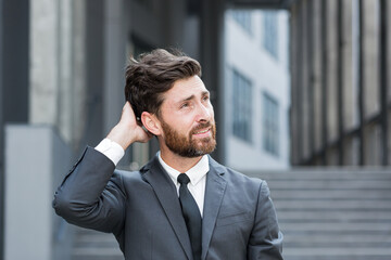 sad worried business bearded caucasian man holding his head. Businessman entrepreneur or employee or worker in suit on city street on background of modern office center. Outside. outdoors
