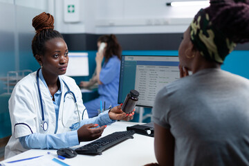 Obraz premium African american physician holding antibiotic bottle in hand discussing disease symptoms with patient explaining medication treatment during clinical consultation. Doctor working in hospital office