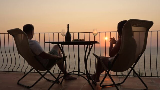 Beautiful young couple drinking wine on the terrace and admiring the sunset over the sea.