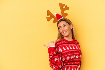 Young caucasian woman wearing a christmas reindeer hat isolated on beige background points with thumb finger away, laughing and carefree.
