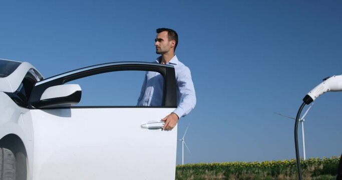 Modern wind turbines generate electricity for ev in sunflower field behind man connecting plug to car at charging station
