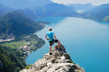 Man with via ferrata gear climbs on top of a rock above Attersee, Austria. Climber, lake, turquoise, summer, tourism, adventure, sport.