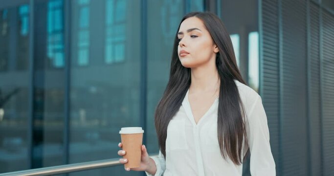 Attractive girl working in an office, corporation, company, dressed in elegant clothes walks in front of a glass building, looks around, coffee break, rest from her duties