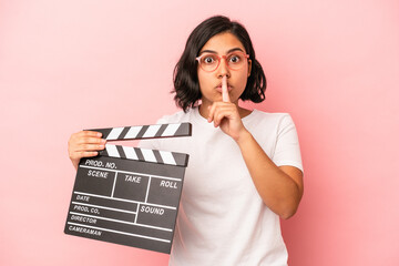 Young latin woman holding clapperboard isolated on pink background keeping a secret or asking for...