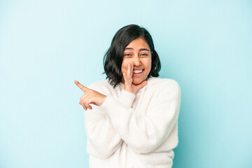 Young latin woman isolated on blue background saying a gossip, pointing to side reporting something.