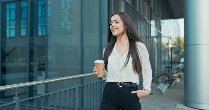 Beautiful cheerful woman working in an office, corporation, company, dressed in elegant clothes walks in front of a glass building, looks around smiling, coffee break, rest from her duties