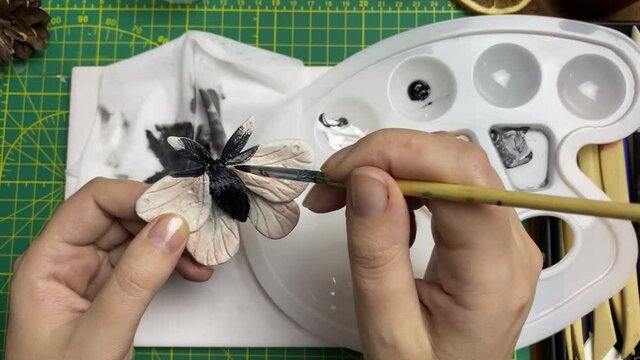 Female's hands paint acrylic brooch in form of butterfly. Palette, brushes, napkins on wooden table. Design of jewelry in form of moth made of clay. Handmade and craft on quarantine.