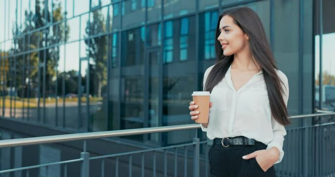 Beautiful cheerful woman working in an office, corporation, company, dressed in elegant clothes walks in front of a glass building, looks around smiling, coffee break, rest from her duties
