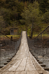 Obraz premium Old wooden suspension bridge across mountain river among autumn forest. Moody image. Countryside background.