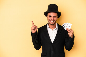 Young caucasian wizard holding magic card isolated on yellow background smiling and pointing aside, showing something at blank space.