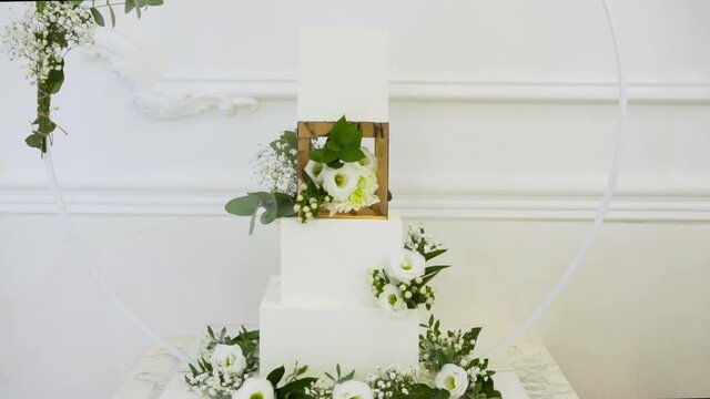 Festive wedding cake decorated with fresh flowers. Sweet confectionery from azyme and cream. The top layers of the cake are separated by a gold cube with flowers. Cake on a round large stand.
