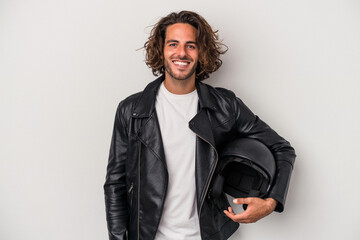 Young biker caucasian man holding a motorbike helmet isolated on gray background happy, smiling and...
