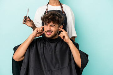 Young arab man ready to get a haircut isolated on blue background covering ears with hands.