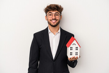 Young arab real estate man holding a model house isolated on isolated background happy, smiling and...
