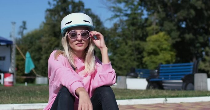 Young blonde woman in protective helmet pink sweatshirt and black skinny trousers sits on curb near cobbled pavement and plays with penny board