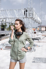 Installation of stage equipment and preparing for a live concert open air. Event manager portrait. Summer music city festival. Young serious woman speaks the walkie-talkie.