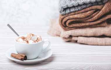 Cozy winter composition, cup with hot chocolate or cocoa with marshmallow and pile of warm knitted clothes on white wooden rustic background. Comfy and soft winter at home, free space for text