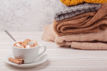 Fototapeta na wymiar Cozy winter composition, cup with hot chocolate or cocoa with marshmallow and pile of warm knitted clothes on white wooden rustic background. Comfy and soft winter at home, free space for text