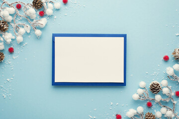 Top view photo of blue envelope with white card christmas decorations snow branches red berries and pine cones on isolated pastel blue background with blank space