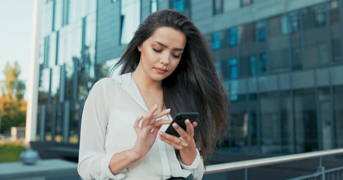Attractive girl working in office corporation, company, dressed in elegant outfit, stands leaning against railing in front of glass building, holding a phone in hand, sending messages, chatting away