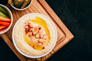 Hummus with olive oil and chickpea in bowl with vegetable sticks and crisp bread on dark stone background.