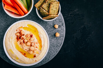 Hummus with olive oil and chickpea in bowl with vegetable sticks and crisp bread on dark stone background.