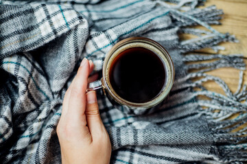 A woman's hand holds a glass mug with hot coffee. A warm plaid scarf with fringe lies on a light...