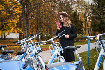 Beautiful young woman in hat holding smartphone and smiling while standing on the street near bicycles. Elegant woman with mobile phone in her hand using bike rental service in autumn park.