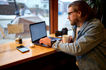 Caucasian young male photographer sitting with laptop and camera at cafe. Concept of modern technology and photoartist. copy space. side view portrait. people lifestyle concept