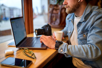 Young successful bearded man sitting at cafe and surfing internet on laptop. Concept of freelance and social networks. copy space. people lifestyle concept. focus on arm with wristwatch