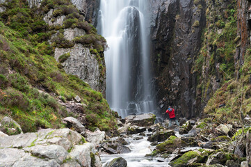 Photographer at Wailing Widows Falls, a famous waterfall in Assynt, North West Highlands of Scotland. On NC500 route. Smooth flow of water, stream, rocks, cliff edges either side with greenery.