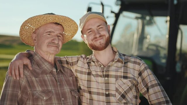 Two farmers are hugging and looking at the camera. They are smiling. There is a tractor behind them. The sun is shining brightly. Portrait shooting. 4K