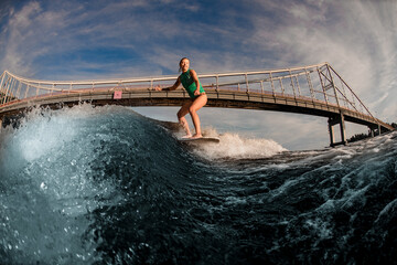 view of beautiful woman wakesurfer riding down on the board on splashing wave against bridge.
