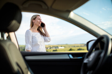 Pretty midle aged woman at the steering wheel of her car, having a break on a long trip, making a phone call
