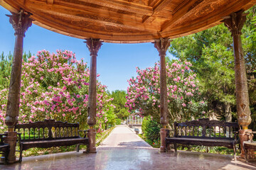Gazebo in Central Asian style with carved columns and benches. Garden and pink bushes of Oleander Nerium on background. Shot in park of historic complex Al-Hakim At Termezi in Termez, Uzbekistan