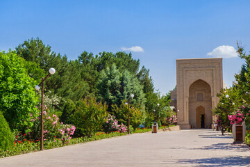 Park alley lushly planted with trees and flowering bushes, leading to main entrance of Al Hakim At-Termezi mosque (its portal is visible). Shot in Termez, Uzbekistan