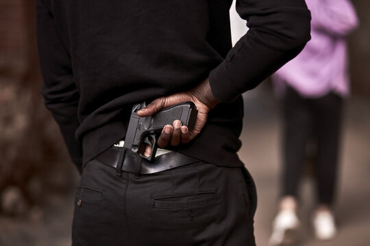 Robbery or criminal man in black hoodie is holding gun standing behind the woman, going to commit a crime. rear view on male in black outfit, close-up hands. criminal activity concept