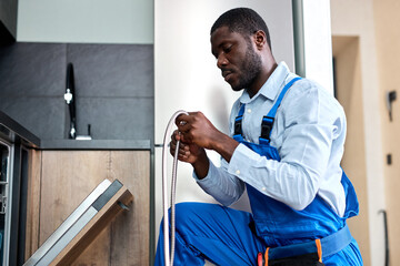 Young Black Handyman Repairing Dishwasher, Need To Change Old Dishwasher Hose, Black Guy In Blue Overalls Workwear Is Concentrated On Work, In Kitchen Indoors. Side View Portrait