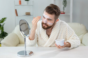 Bearded guy applying facial cream in front of mirror Skin care and spa for man concept.