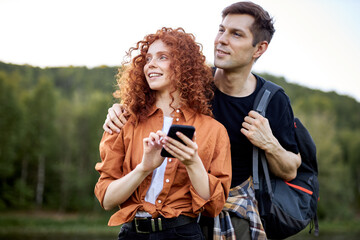 Smiling caucasian couple redhead woman and man travelers standing by river using smartphone, looking at online map, tourists in casual wear traveling together. travel, trip and vacation concept