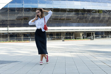 A young woman in glasses, a white shirt and a black skirt is talking on the phone against on the street