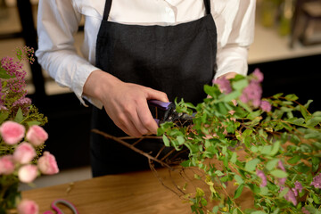 cropped female holding scissors while cutting flowers, preparing bouquet in flower shop. unrecognizable woman in black apron is arranging wild flowers petals, preparing them for decoration