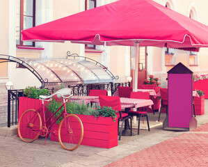 A flowerbed made from a bicycle and tubs of flowers in the summer caf&eacute;. Stone sidewalk, tables, tablecloths, chairs, protective awning. Photo.