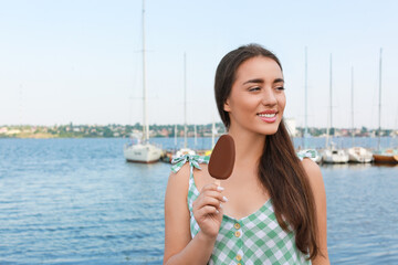 Beautiful young woman holding ice cream glazed in chocolate near river, space for text
