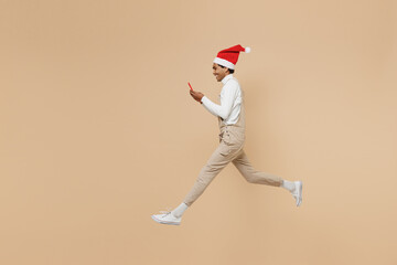 Fototapeta na wymiar Full body side view young african man 20s wear Santa Claus red Christmas hat jump high use mobile cell phone isolated on plain pastel beige background studio. Happy New Year 2022 celebration concept.