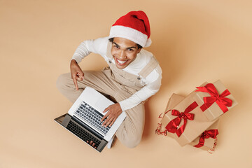 Full body overview young african man 20s wearing Santa Claus red Christmas hat sit near present gifts hold use point on laptop pc computer isolated on plain pastel beige background studio portrait