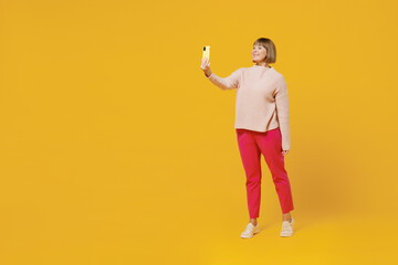 Fototapeta na wymiar Elderly fun woman 50s wears pink casual knitted sweater do selfie shot on mobile cell phone post photo on social network isolated on plain yellow background studio portrait. People lifestyle concept.