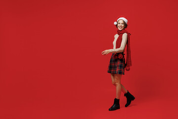 Full body side view young smiling caucasian woman 20s wear Santa Claus Christmas red hat walk going strolling isolated on plain red background studio portrait. Happy New Year 2022 celebration concept.